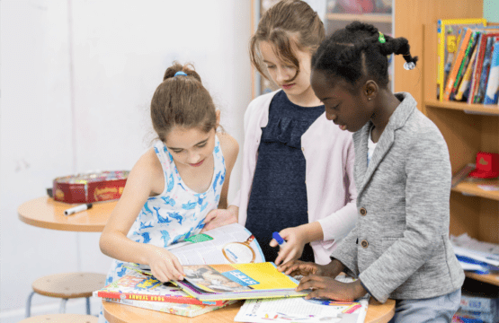 3 young girls discover more standing around a table looking at books together.